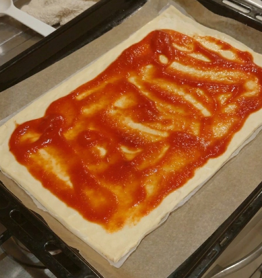 Ein rechteckiger Pizzaboden mit gleichmäßig verteilter Tomatensauce, wobei ein kleiner Rand freigelassen wurde.