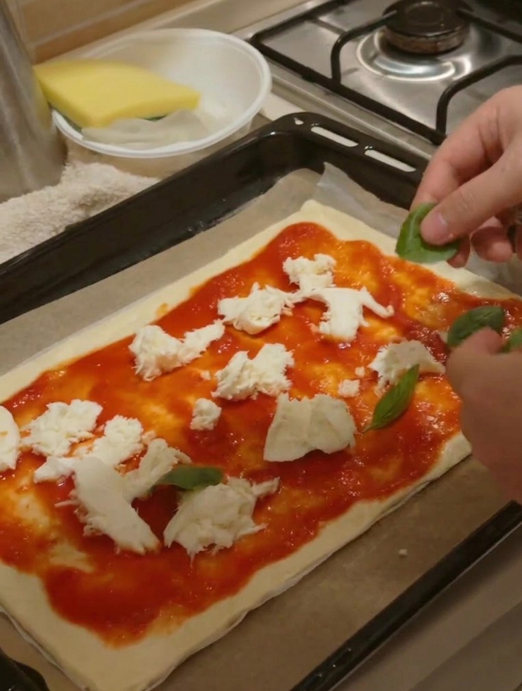 Hände legen frische grüne Basilikumblätter auf eine Pizza, die bereits mit Sauce und Käse belegt ist.