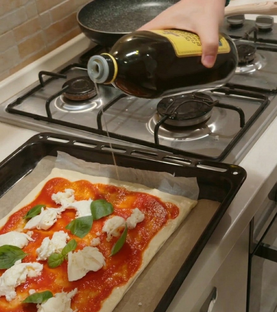 Eine Hand gießt einen Strahl Olivenöl aus einer Glasflasche auf einen rohen Pizzaboden mit Tomatensauce, Mozzarella und Basilikum.