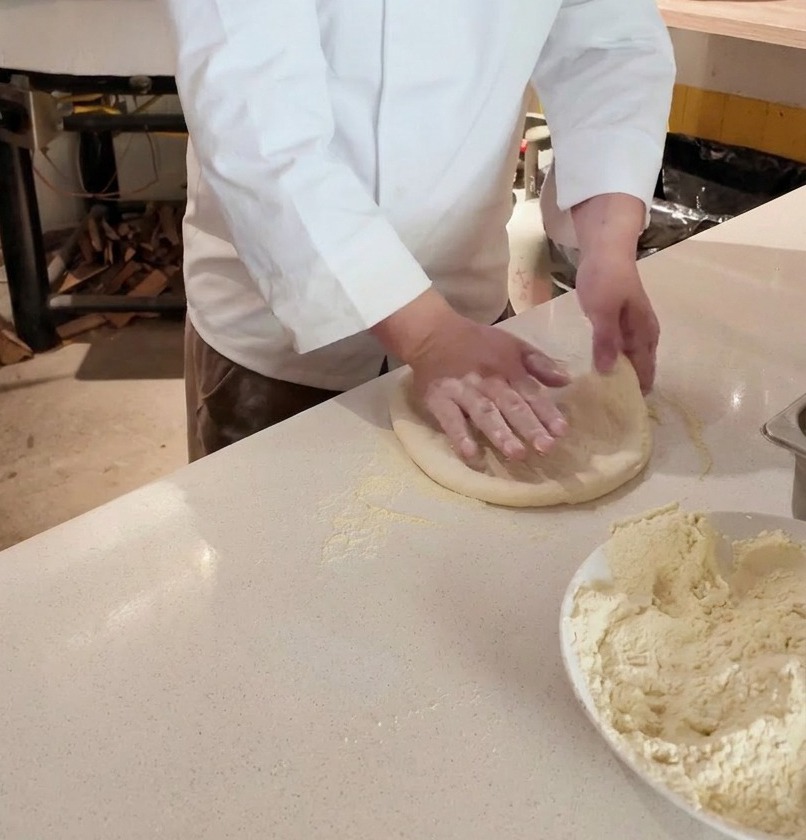 Hände eines Kochs dehnen ein rundes Stück rohen Pizzateig auf einer bemehlten Arbeitsfläche vor einem Holzofen.