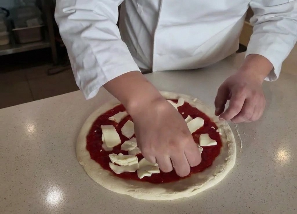 Hände platzieren gezupfte Stücke weißen Mozzarella auf einer Pizza mit roter Tomatensauce.