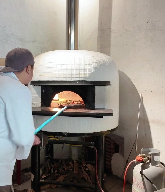 Eine Pizza wird von einem langen Pizzaschieber in die Öffnung eines glühenden Steinofens geschoben.