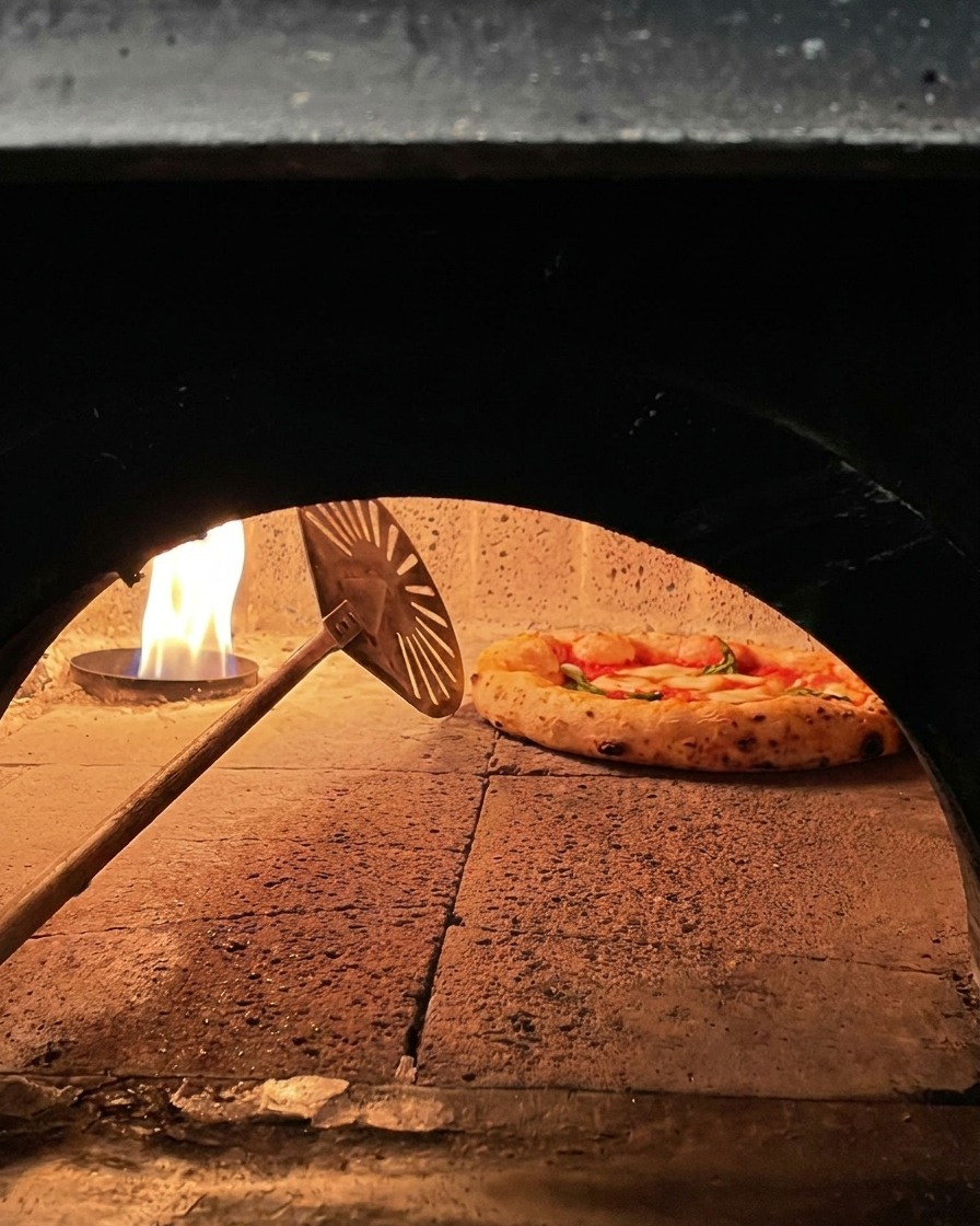 Eine Pizza Margherita wird im Holzofen mit einem kleinen Metallschieber nahe am Feuer gedreht.