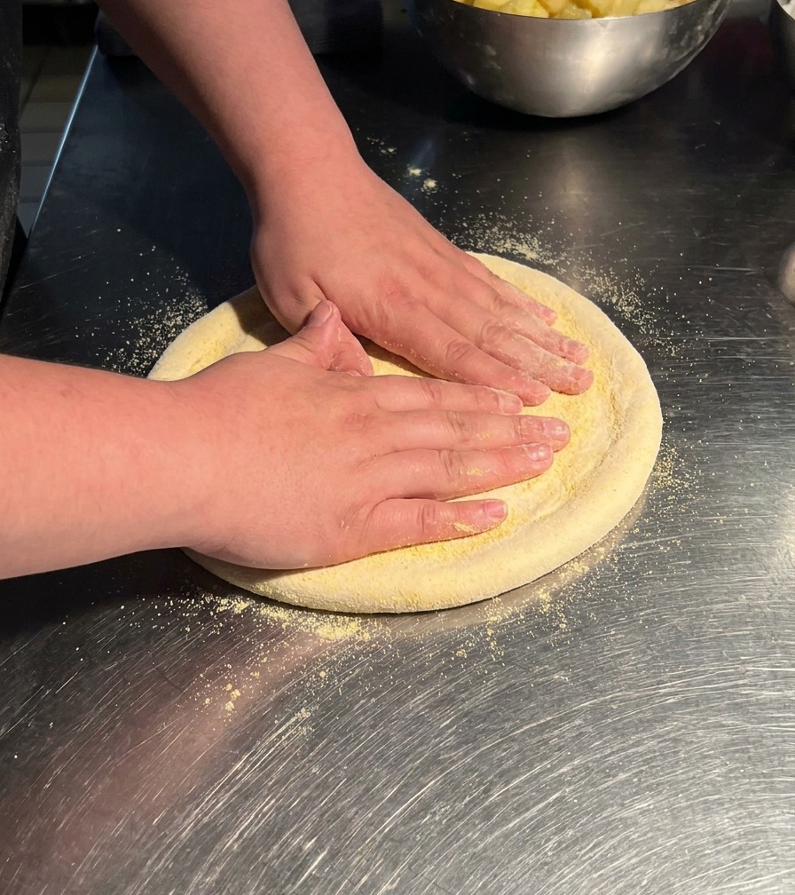 Hände drücken ein kreisförmiges Stück Pizzateig auf einem Edelstahltisch flach und dehnen es zu einer größeren Scheibe aus.