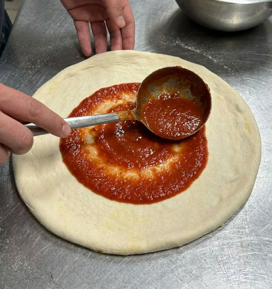 Eine Metallkelle verteilt leuchtend rote Tomatensauce in kreisenden Bewegungen auf dem runden Pizzateig.