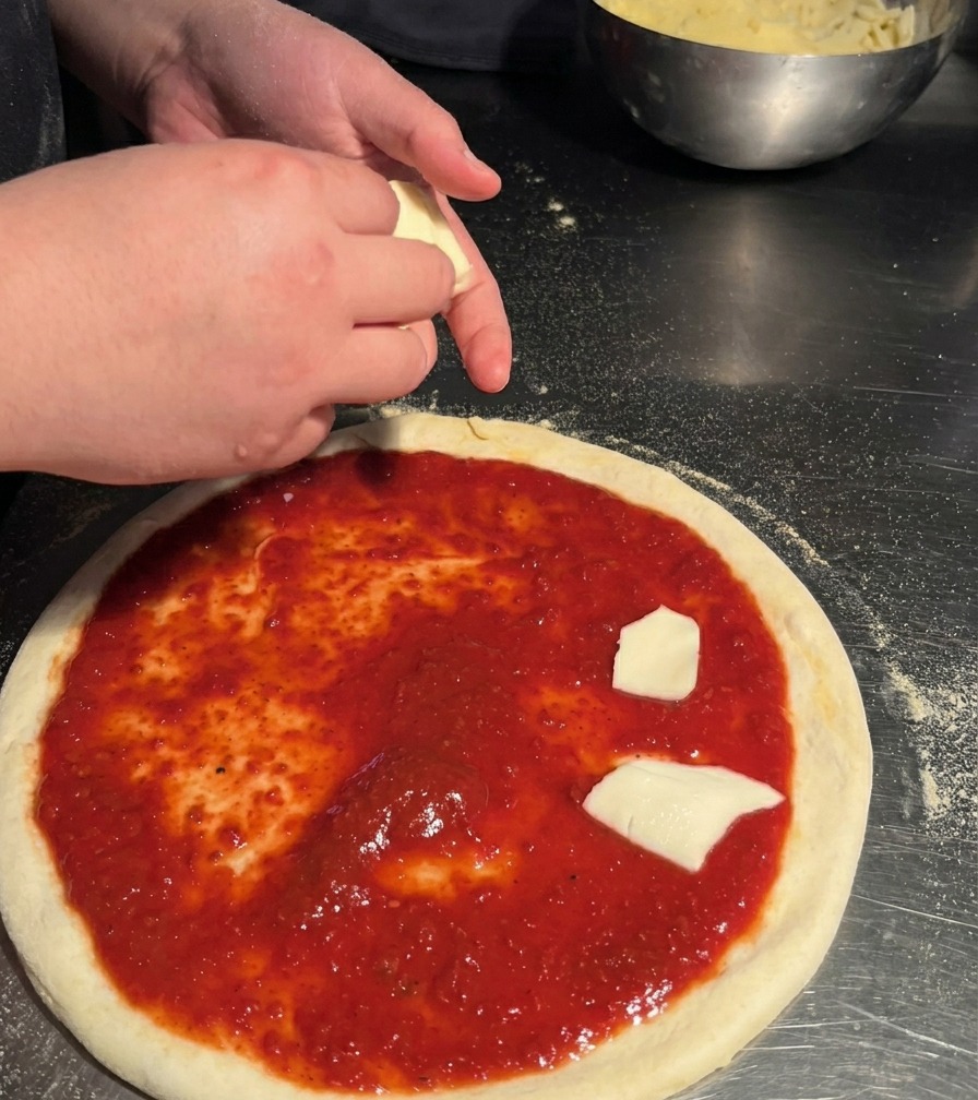 Eine Person verteilt von Hand gezupfte Stücke weißen Mozzarellas auf dem mit Sauce bestrichenen Pizzaboden.
