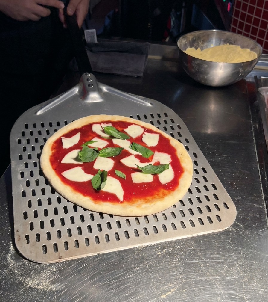 Eine rohe Pizza Margherita liegt fertig belegt auf einem perforierten Pizzaschieber aus Metall.