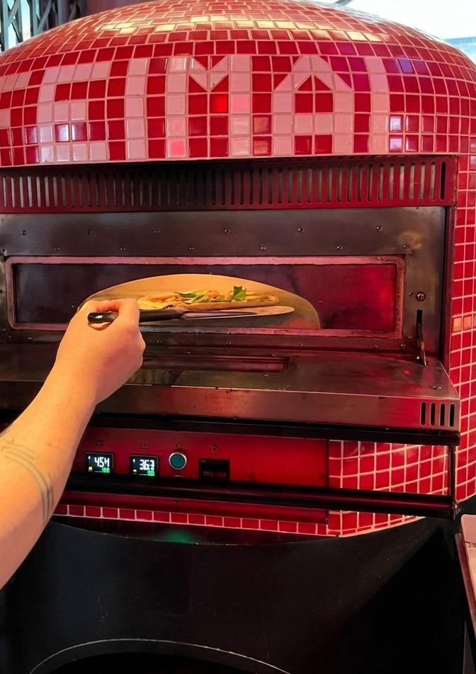 Eine Person schiebt eine rohe Pizza von einem Metallschieber in die glühende Öffnung eines traditionellen Pizzaofens.