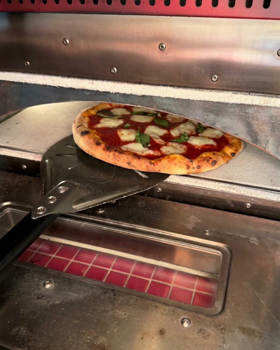 Eine frisch gebackene Pizza mit knusprigem Rand wird mit einem Metallschieber aus dem Ofen geholt.