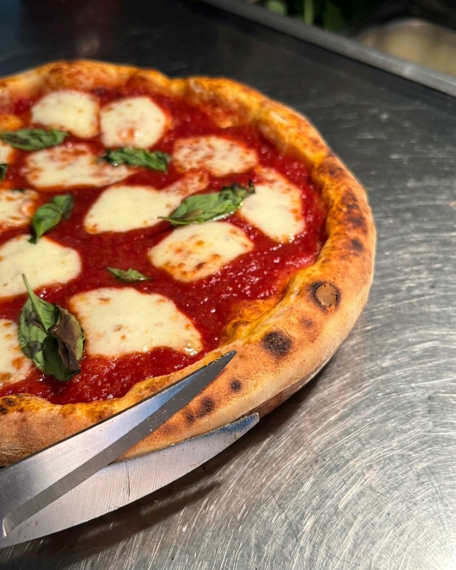 Nahaufnahme einer Metallschere, die durch den fluffigen Rand einer heißen Pizza schneidet.