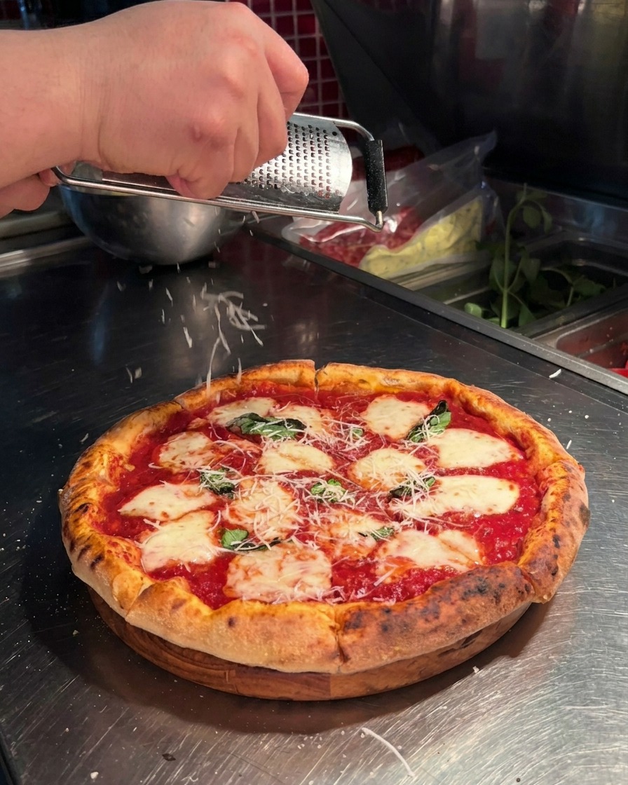 Nahaufnahme einer Hand, die mit einer Microplane-Reibe frischen Parmesan über die Pizza reibt.
