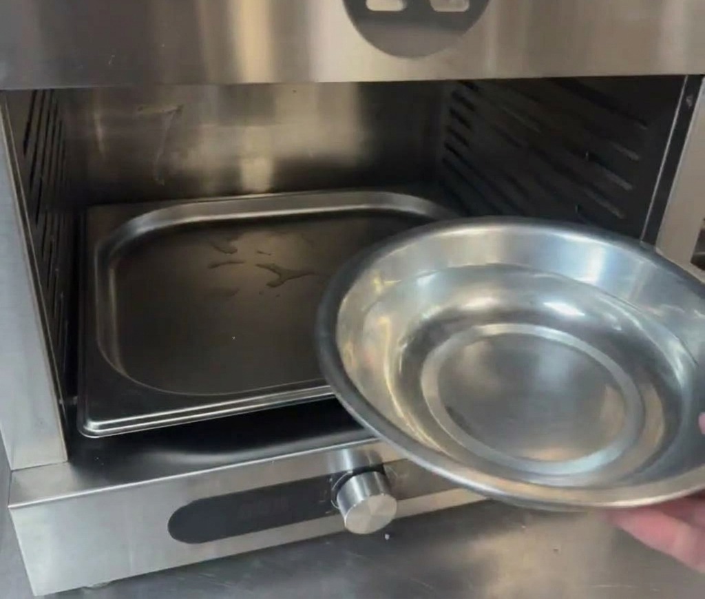 Eine Hand gießt Wasser aus einer kleinen Metallschale in eine Schale am Boden eines Profi-Ofens.