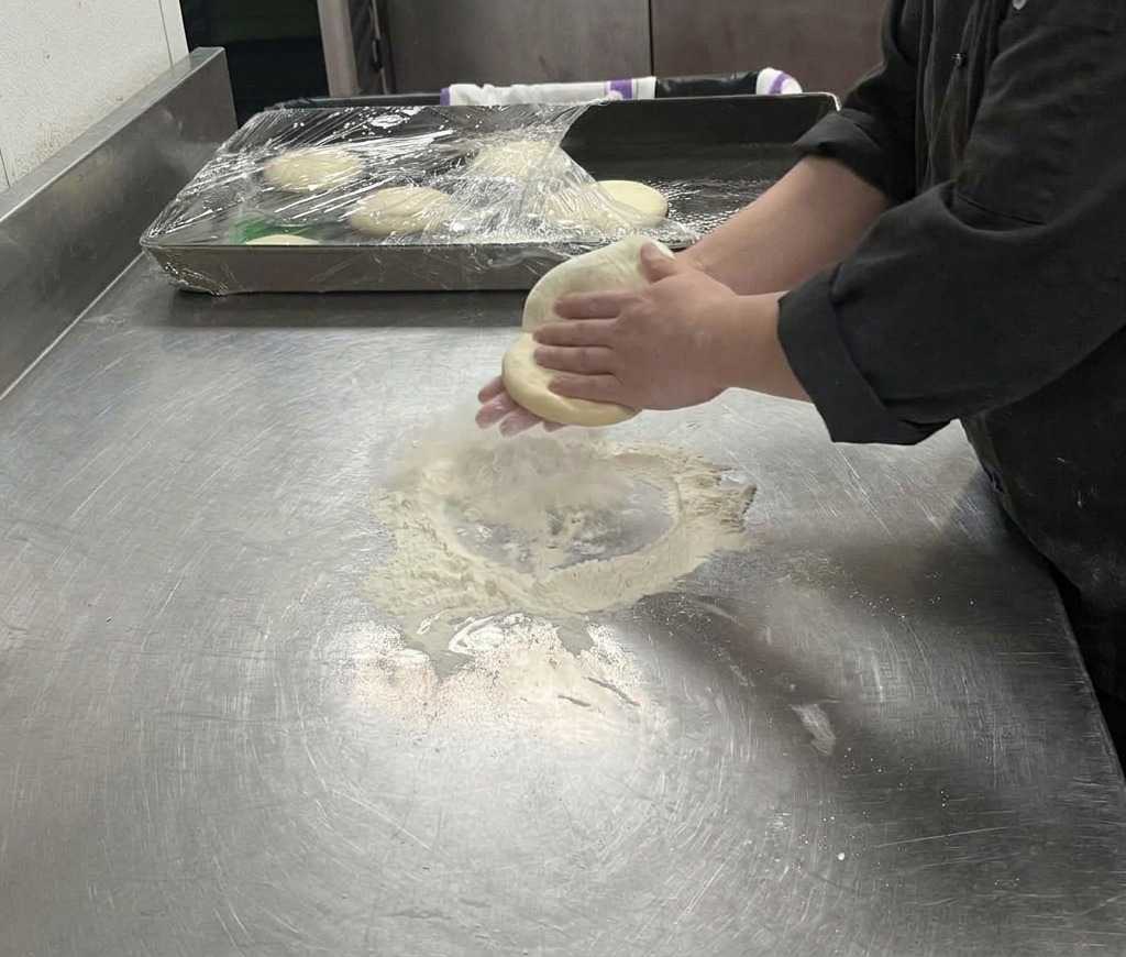Hände dehnen einen hellen Pizzateig über einer bemehlten Fläche zu einem größeren Kreis.