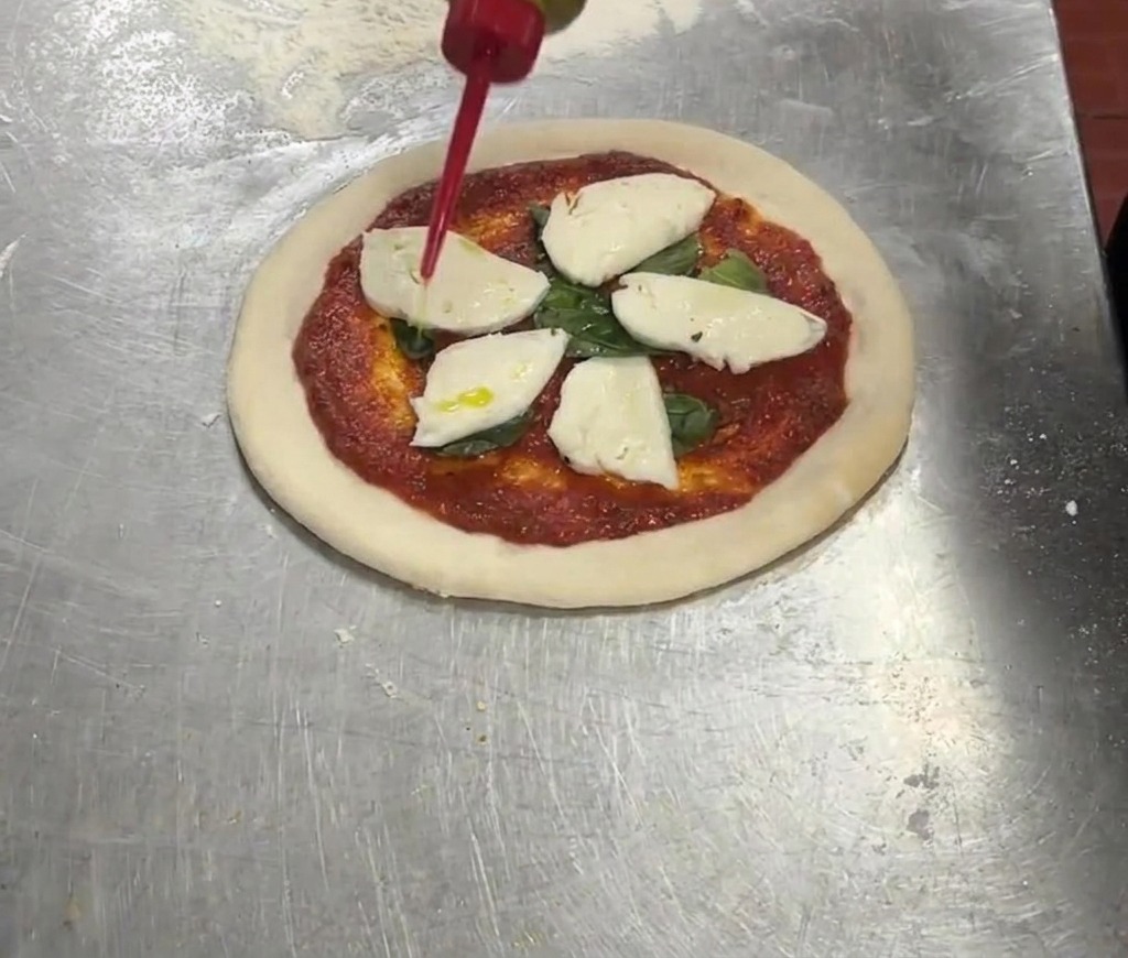 Eine rote Spritzflasche beträufelt eine Pizza mit Olivenöl nativ extra.