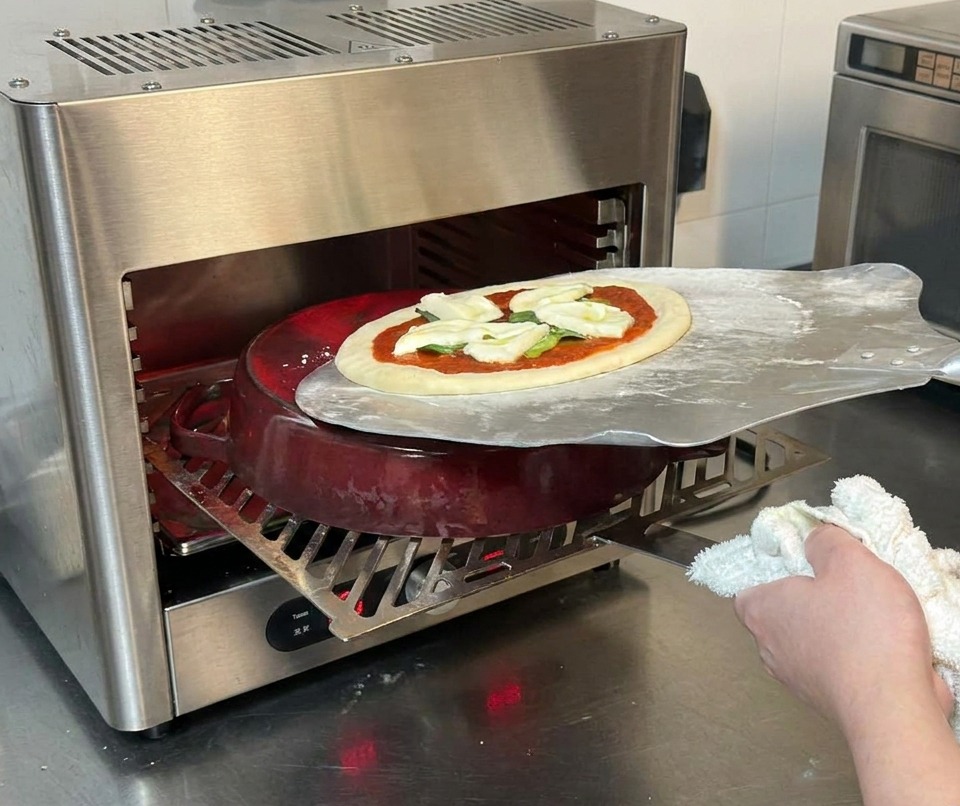 Ein Pizzaschieber aus Metall gleitet eine rohe Pizza auf einen roten Gusseisentopf in einem Ofen.