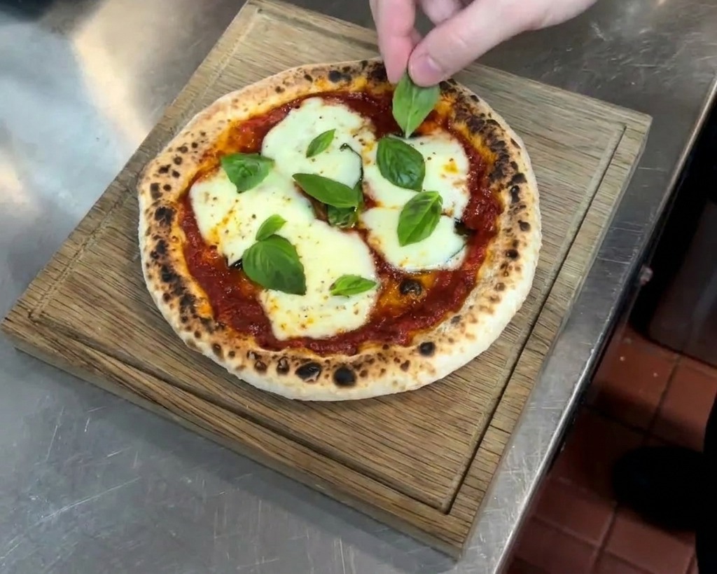 Eine Hand legt frische Basilikumblätter auf eine heiße Pizza mit geschmolzenem Käse und dunklen Röstpunkten.