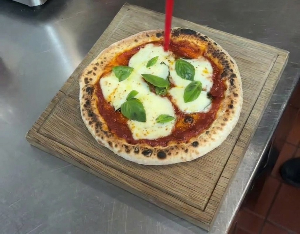 Ein Strahl Olivenöl wird aus einer Spritzflasche auf eine fertige Pizza Margherita gegossen.