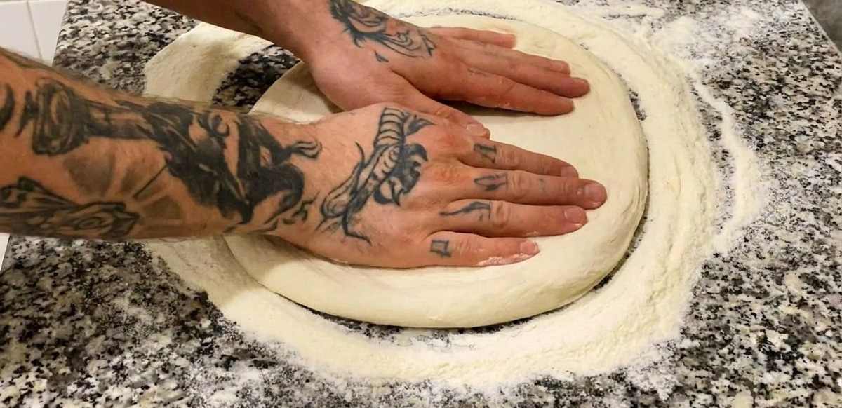 Zwei Hände drücken auf einer bemehlten Marmorplatte einen runden Pizzateig flach.