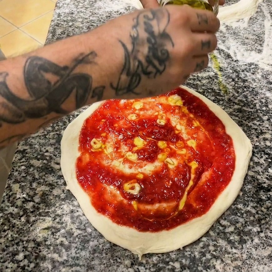 Eine Hand verteilt mit einem Löffel leuchtend rote Tomatensauce spiralförmig auf dem rohen Pizzateig.