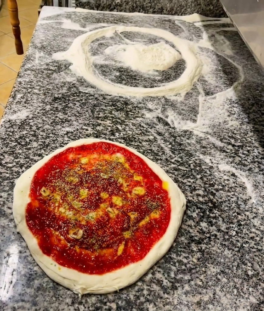Eine Hand streut getrocknete Kräuter über den mit Sauce und Öl bestrichenen Pizzaboden.