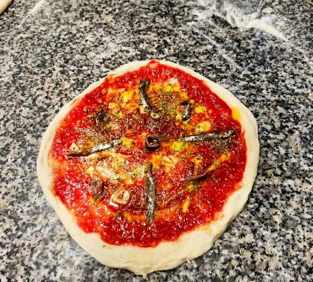 Draufsicht auf eine Pizza mit Tomatensauce und Kräutern, auf der Sardellenfilets wie Speichen eines Rades angeordnet sind.