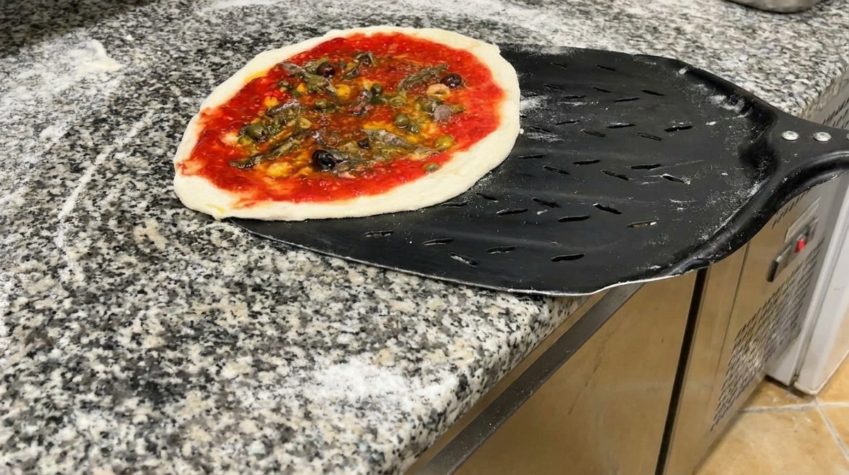 Eine fertig belegte Pizza liegt auf einem perforierten schwarzen Metallschieber bereit zum Backen.