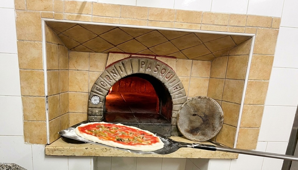Eine Pizza wird mit einem Schieber in einen traditionellen Steinofen mit lodernden Flammen geschoben.