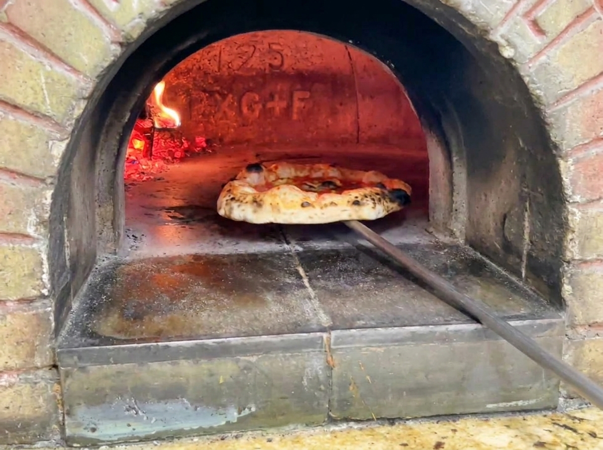 Die fertige Pizza wird aus dem Ofen geholt; sie zeigt einen goldbraunen, blasigen Rand mit dunklen Röstpunkten.