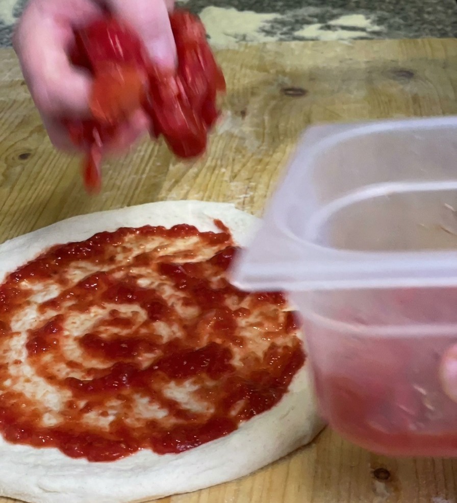 Eine Hand lässt Stücke von dickflüssigem Tomatenfruchtfleisch auf den bereits mit Sauce bestrichenen Pizzaboden fallen.