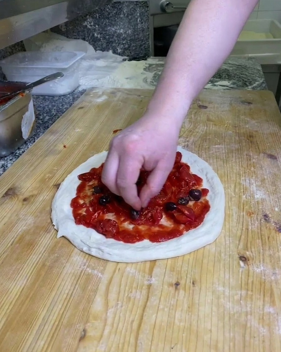 Eine Hand platziert dunkle, ganze Oliven auf dem tomatenbedeckten Pizzateig auf einer bemehlten Arbeitsfläche.