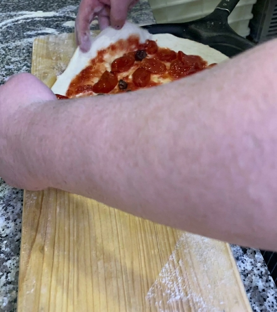 Der Koch schiebt eine rohe, belegte Pizza von einer Holzwerkbank auf einen schwarzen Metall-Pizzaschieber.