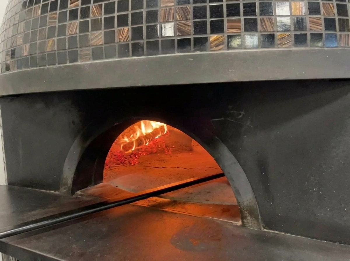 Blick in die glühende Öffnung eines traditionellen Pizzaofens mit Flammen unter der Kuppel und einer backenden Pizza.