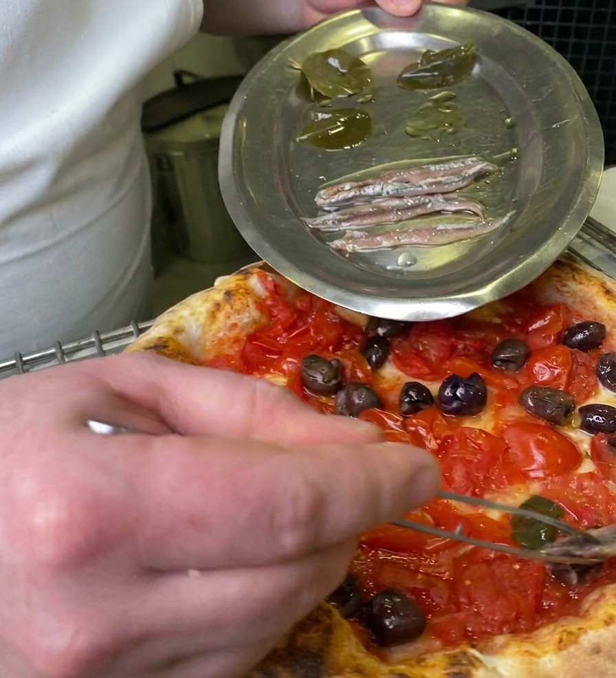 Ein Koch nutzt eine Pinzette, um ein eingelegtes Lorbeerblatt von einem Metallteller auf die Pizza zu legen.