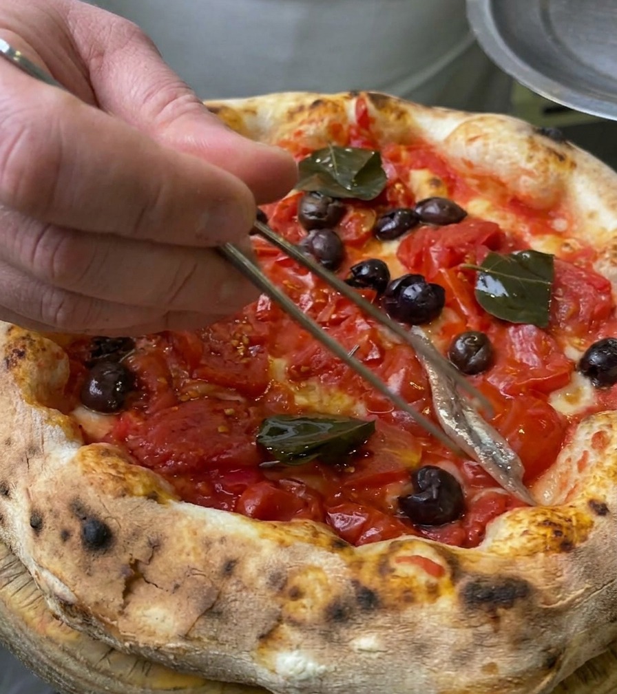 Nahaufnahme eines silbernen Sardellenfilets, das mit einer Küchenpinzette auf den Pizzarand gelegt wird.