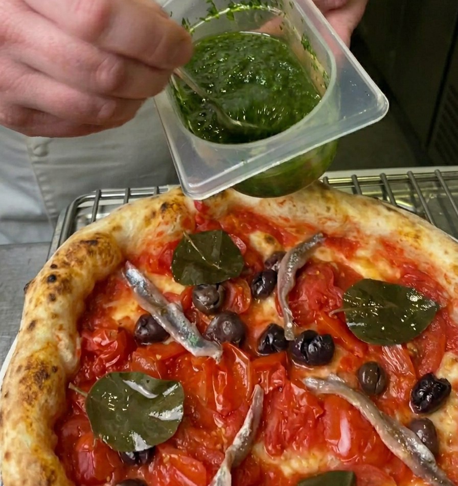 Ein Koch träufelt eine leuchtend grüne Kräutersauce mit einem kleinen Löffel aus einem Behälter über die Pizza.