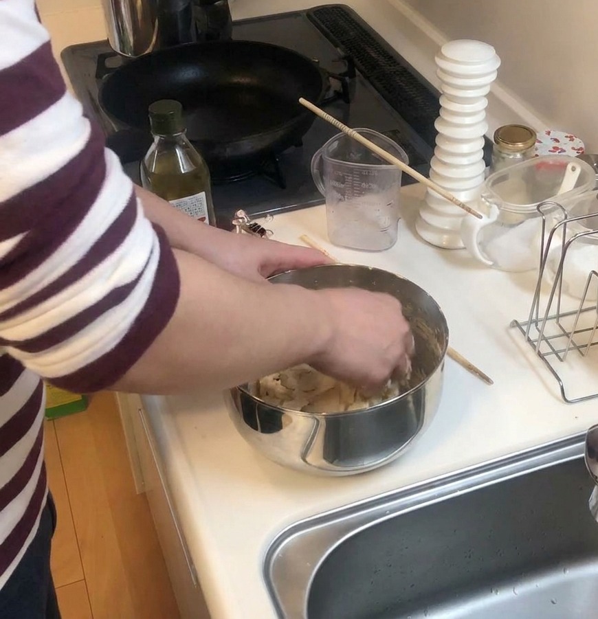 Hände kneten Pizzateig in einer Edelstahlschüssel auf einer weißen Arbeitsplatte neben einer Flasche Olivenöl.