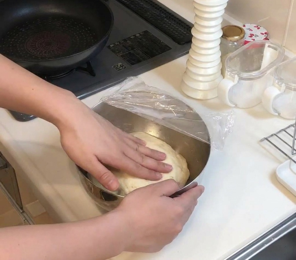 Hände drücken sanft auf einen großen Kloß aufgegangenen Teigs in einer mit Frischhaltefolie abgedeckten Schüssel.