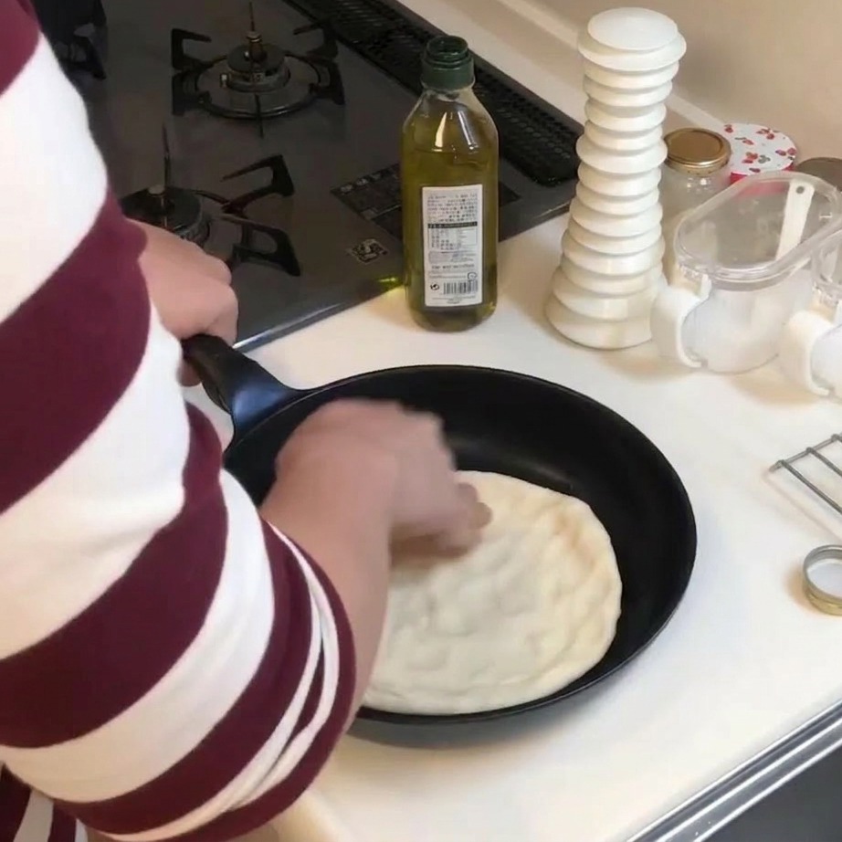 Eine Person drückt mit den Händen einen Kreis aus Pizzateig flach in eine schwarze Bratpfanne.