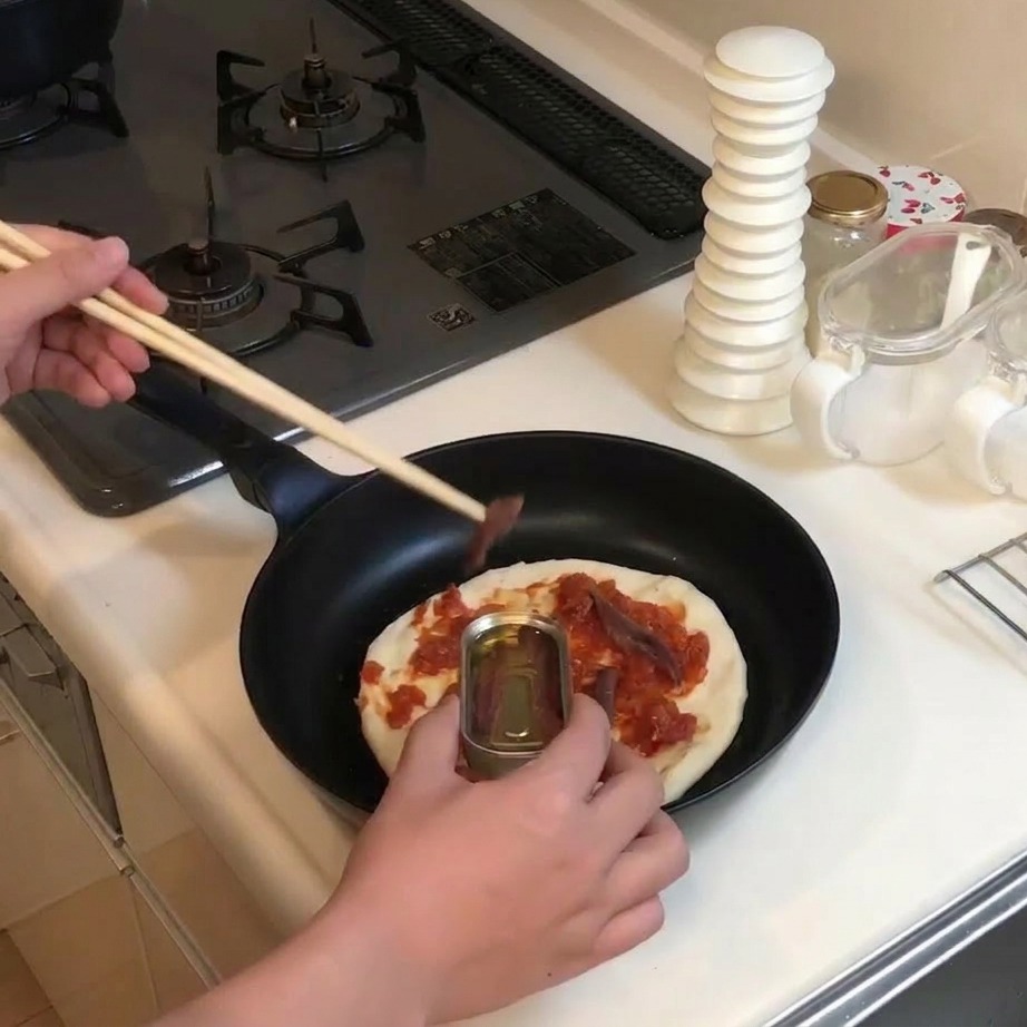 Mit Holzstäbchen werden Sardellen aus einer kleinen Metalldose auf die mit Sauce bestrichene Pizza gelegt.