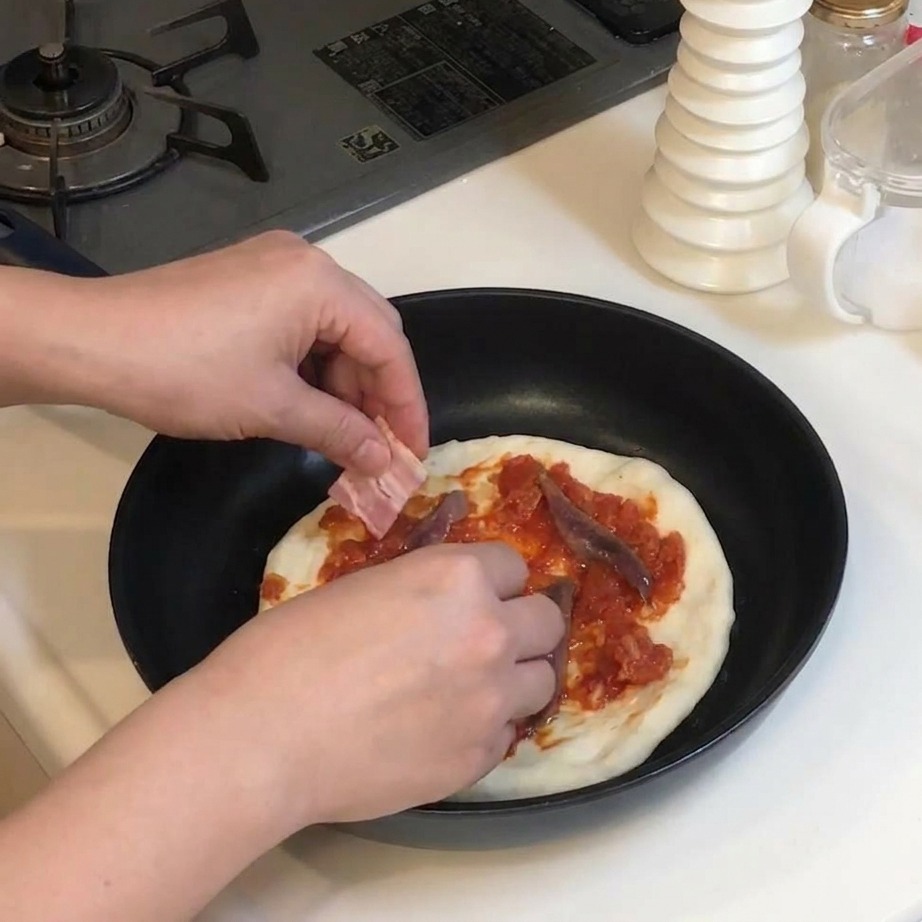 Hände legen rechteckige Streifen von rohem Speck auf eine Pizza, die bereits mit Sauce und Sardellen belegt ist.