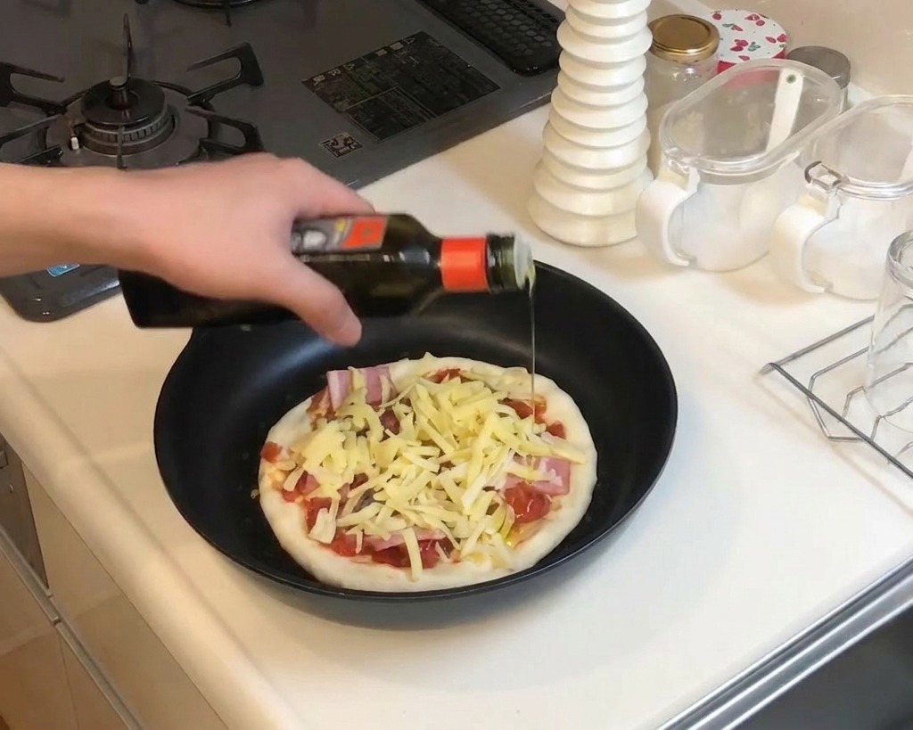Nahaufnahme einer Hand, die Olivenöl aus einer dunklen Flasche über die vorbereitete Pizza in der Pfanne träufelt.
