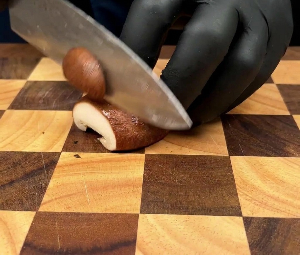 Nahaufnahme einer Person mit schwarzen Handschuhen, die einen Kräuterseitling auf einem Holzbrett in dünne Scheiben schneidet.