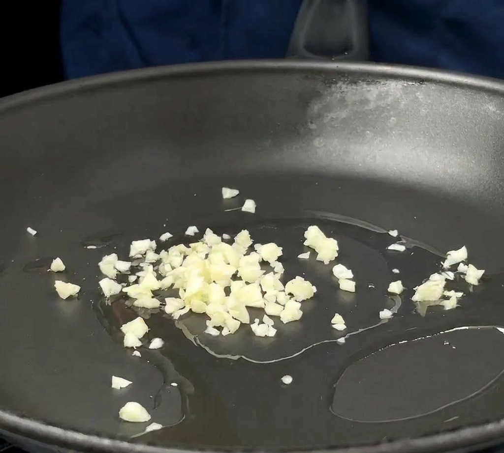 Gehackter Knoblauch und rote Zwiebeln brutzeln in etwas Öl in einer dunklen Pfanne.