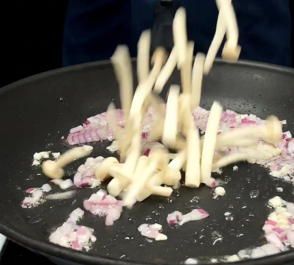 Weiße Buchenpilze werden in eine Pfanne zu den sautierten Zwiebeln und dem Knoblauch gegeben.
