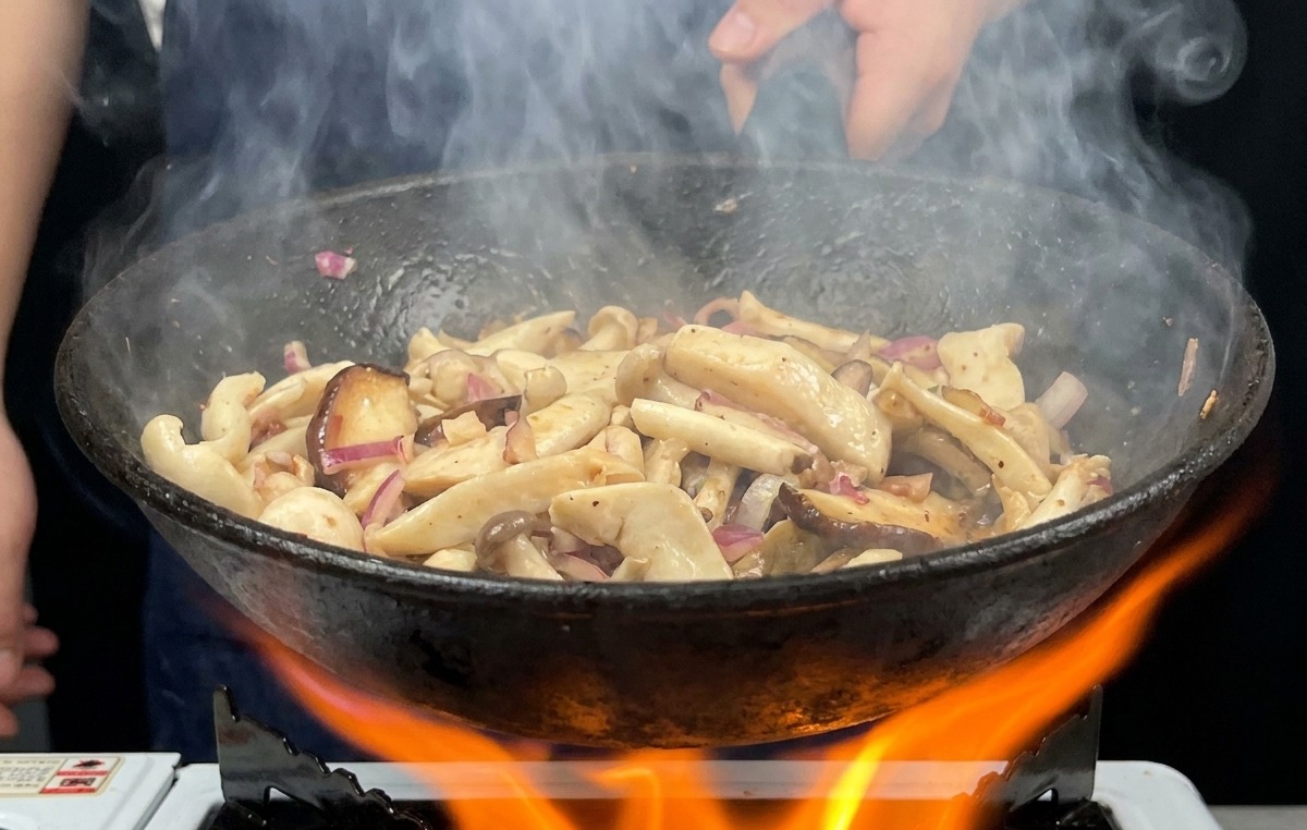Eine Pfanne mit Pilzen wird über einer hellorangen Gasflamme geschwenkt, Dampf steigt auf.