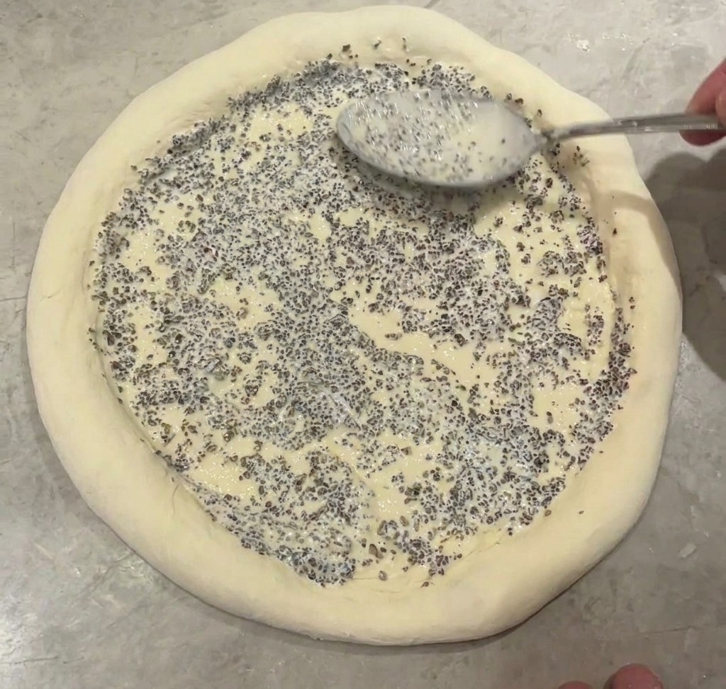 Ein Metalllöffel verteilt eine dickflüssige, grau gesprenkelte Trüffelsauce in der Mitte des Pizzabodens.