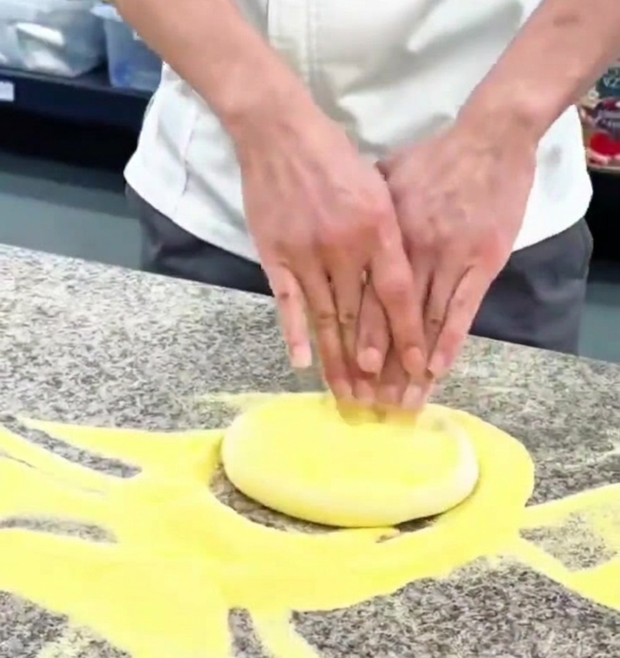 Nahaufnahme von Händen, die eine runde Kugel Pizzateig auf einer bemehlten Granitfläche flach drücken.