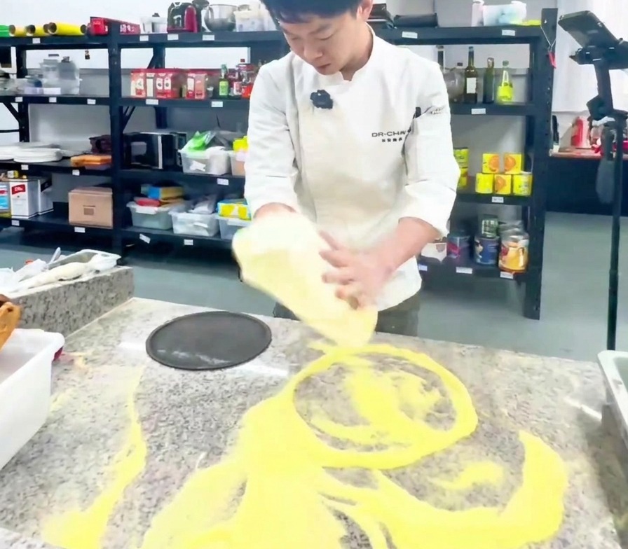 Ein Koch in weißer Uniform dehnt einen großen Pizzateig vertikal über den Handrücken in einer Profiküche.