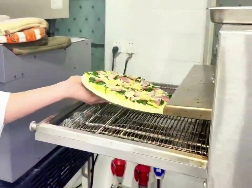 Ein Mitarbeiter schiebt die rohe Spinat-Speck-Pizza auf das Metallband eines professionellen Pizzaofens.