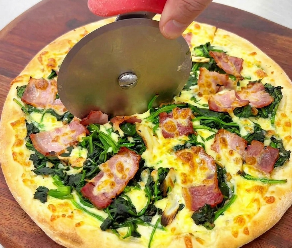 Nahaufnahme eines Pizzaschneiders, der durch eine goldbraun gebackene Pizza mit Spinat und Speck gleitet.